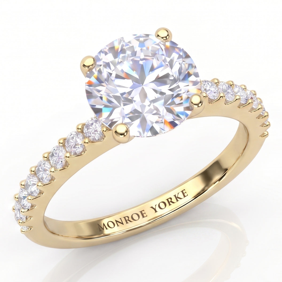 Spring - a yellow gold engagement ring featuring a large round brilliant-cut center stone in a 4-claw setting and a pavé band with small accent stones on the b and. The brand name 'MONROE YORKE' is engraved on the inner band.
