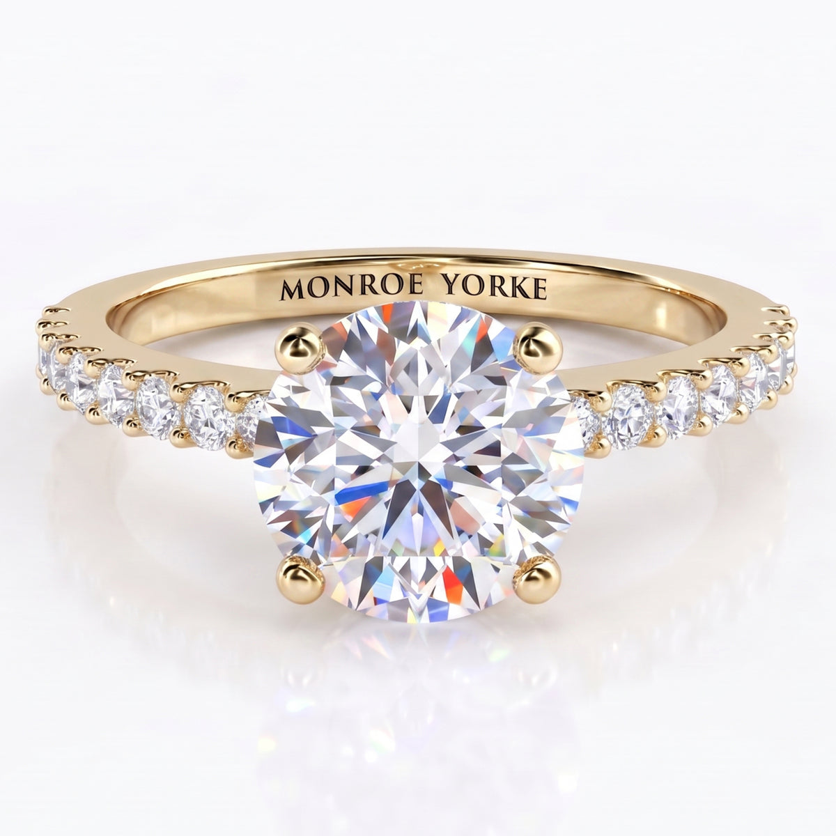 Spring - a yellow gold solitaire engagement ring with a round brilliant-cut center stone in a 4-claw setting and small pavé-set accent stones along the shoulders, engraved with the brand name 'MONROE YORKE' on the band.