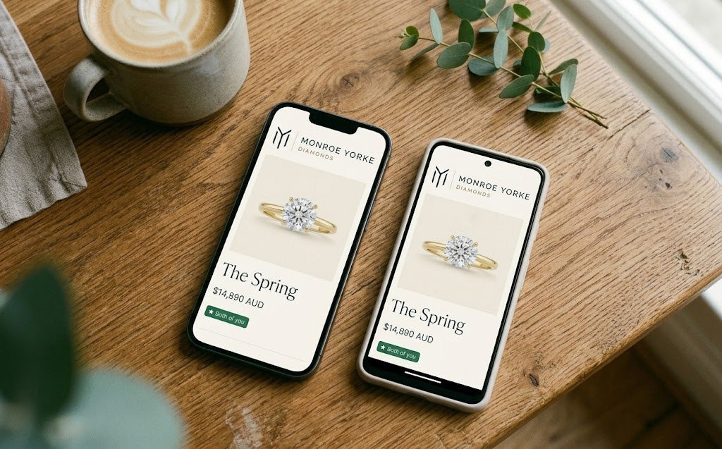 Two phones on a wooden kitchen table, both showing the same Monroe Yorke engagement ring page — coffee cup on the left, eucalyptus sprig on the right, late morning light