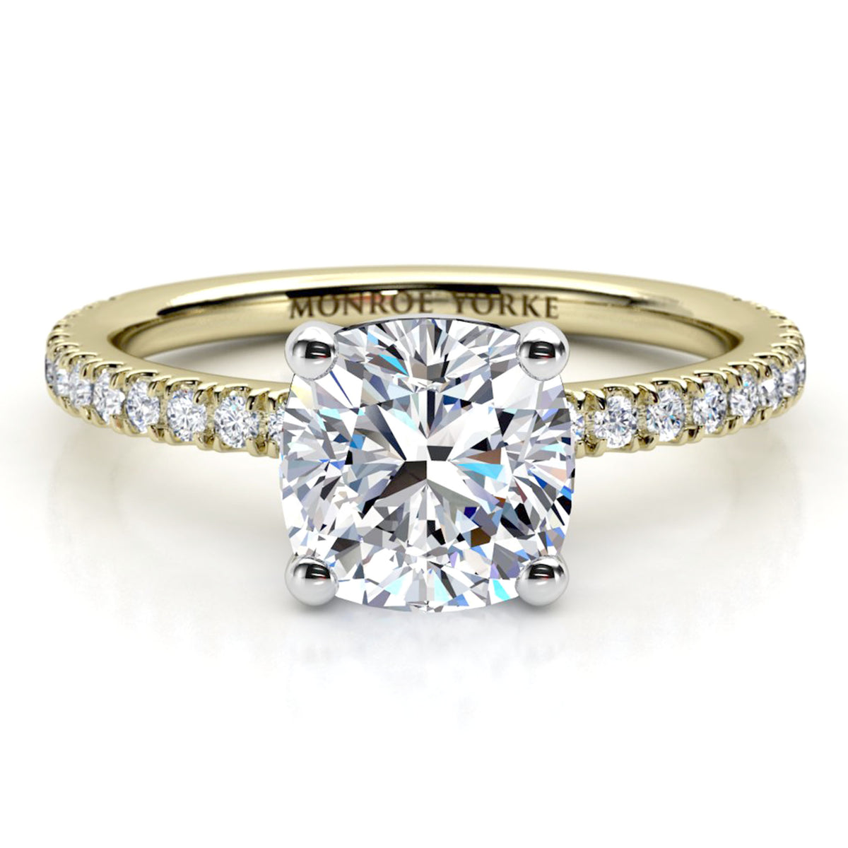 A yellow gold solitaire engagement ring featuring a cushion cut diamond center stone and a pavé-set diamond band. The ring is photographed on a clean white background with a soft reflection.