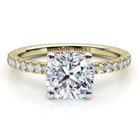 A yellow gold solitaire engagement ring featuring a cushion cut diamond center stone and a pavé-set diamond band. The ring is photographed on a clean white background with a soft reflection.