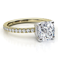 A yellow gold solitaire engagement ring with a cushion-cut diamond center stone and a row of small round diamonds on the band, shown on a white background. 