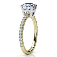 A yellow gold engagement ring featuring a cushion cut center stone and a pavé-set diamond band, shown on a clean white background.