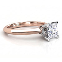 A close-up studio shot of a classic princess cut diamond solitaire engagement ring. The ring features a rose gold band with a polished white metal four-prong setting holding the diamond.