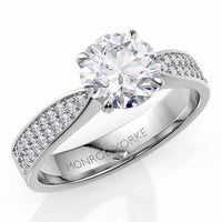 A white gold or platinum diamond engagement ring with a round-cut center stone and pavé-set side stones, shown on a clean white background. 