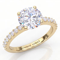 Spring - a yellow gold engagement ring featuring a large round brilliant-cut center stone in a 4-claw setting and a pavé band with small accent stones on the b and. The brand name 'MONROE YORKE' is engraved on the inner band.