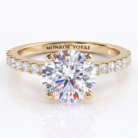 Spring  - a yellow gold solitaire engagement ring with a round brilliant-cut center stone in a 4-claw setting and small pavé-set accent stones along the shoulders, engraved with the brand name 'MONROE YORKE' on the band.