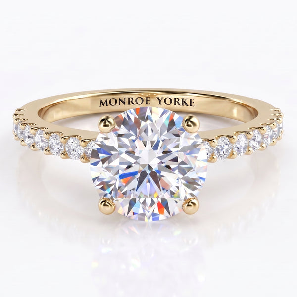 Spring  - a yellow gold solitaire engagement ring with a round brilliant-cut center stone in a 4-claw setting and small pavé-set accent stones along the shoulders, engraved with the brand name 'MONROE YORKE' on the band.